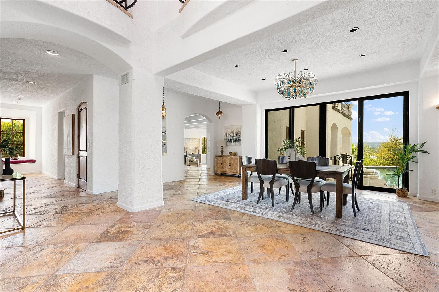 This interior shot showcases a dining room with travertine flooring and white textured walls. A wooden dining table with upholstered chairs sits atop a patterned area rug, illuminated by a decorative chandelier. Large sliding glass doors offer a view of the exterior, blending indoor and outdoor living.