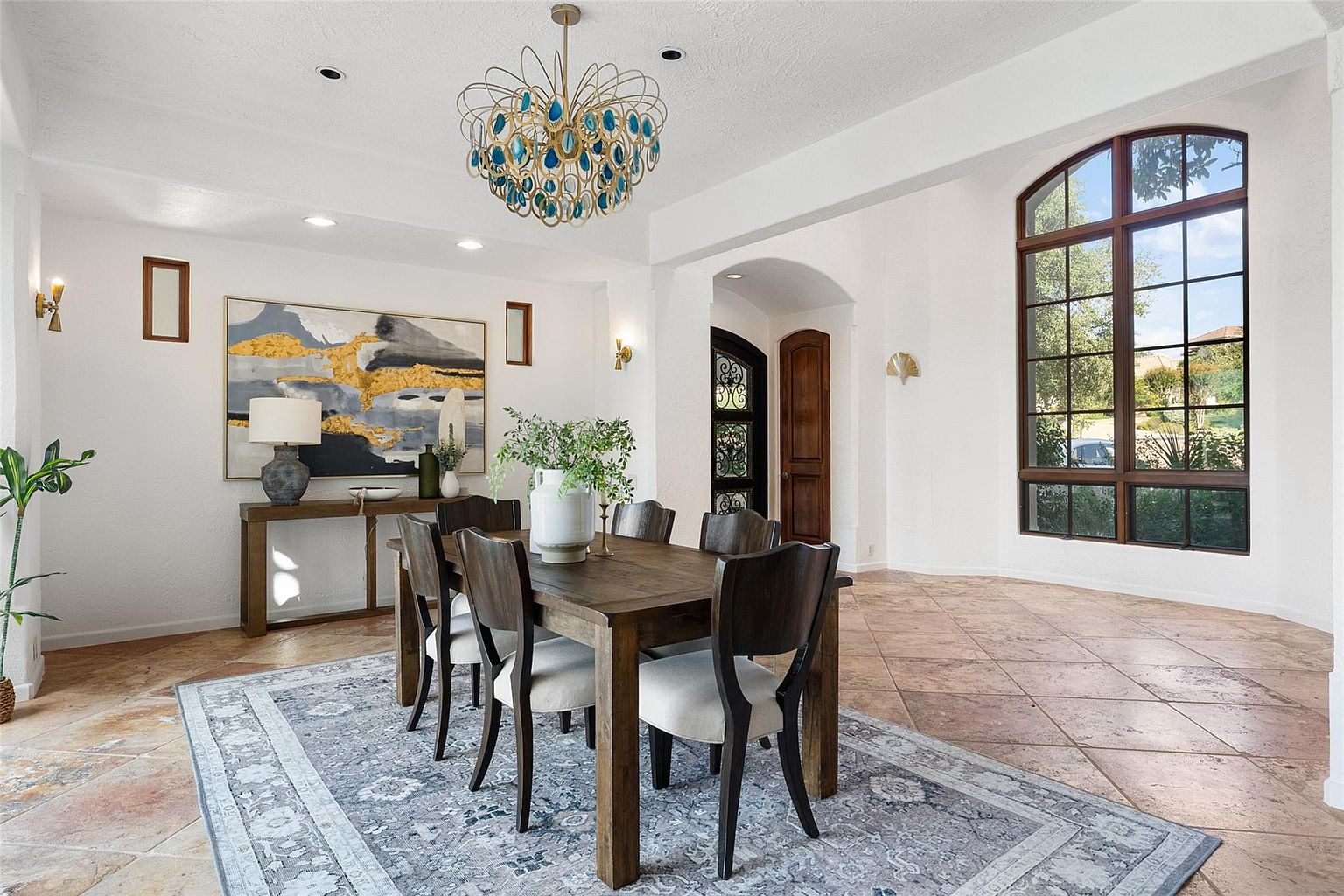 This is an interior shot of a dining room featuring a wooden dining table with upholstered chairs, set on a patterned area rug. A large abstract painting hangs on the wall above a console table with a lamp and decorative objects. The room is well-lit with a decorative chandelier and natural light from a large arched window, creating a bright and inviting atmosphere.