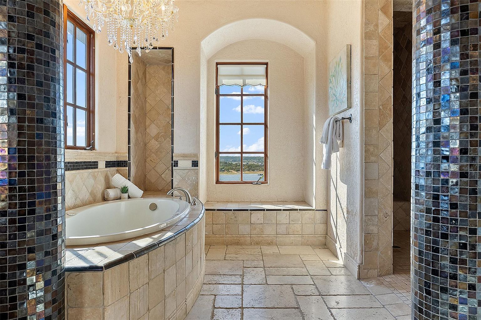 This is a luxurious primary bathroom featuring a large soaking tub with tile surround, set beneath a window that offers a view of the outdoors. The bathroom has travertine tile flooring and mosaic tile accents on the columns and walls. A chandelier hangs above the tub, adding to the opulent feel of the space.