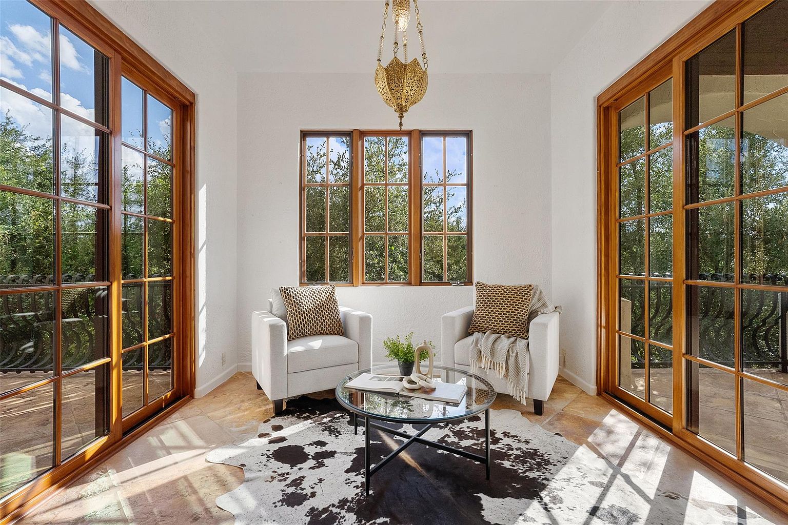 This sunroom features two white armchairs with patterned pillows, flanking a round glass-topped coffee table on a cowhide rug. Natural light floods the space through large windows and doors with wooden frames, offering views of the surrounding greenery. An ornate chandelier hangs from the ceiling, adding a touch of elegance.