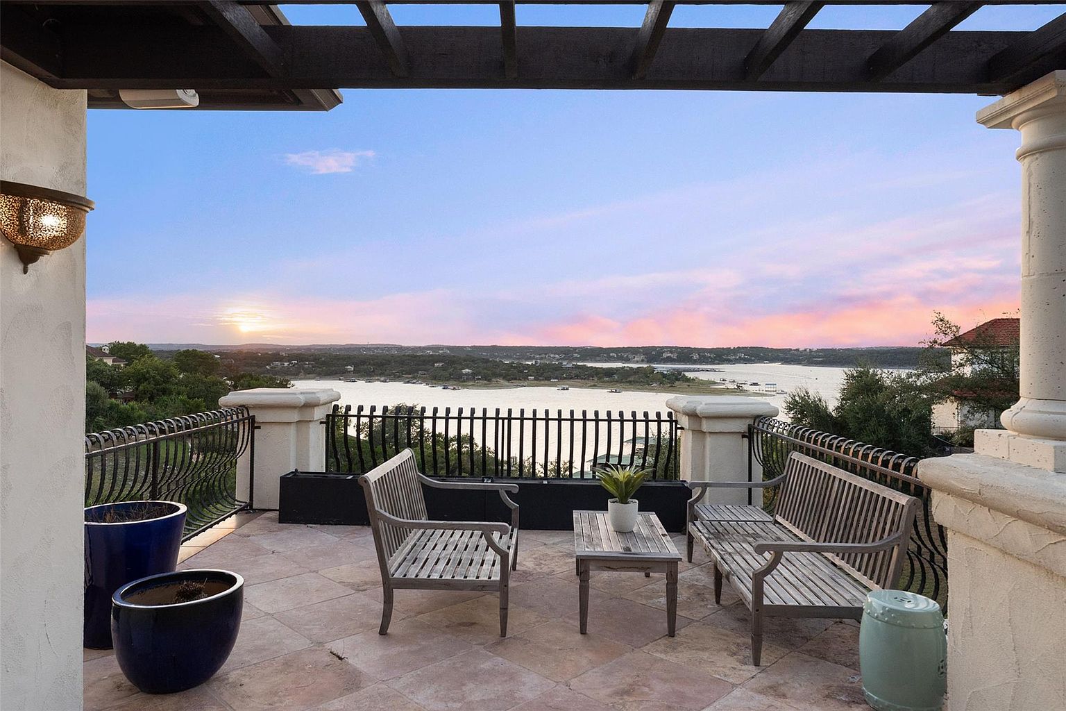 This image showcases a serene balcony or patio with a stunning view of a lake at sunset. The space is furnished with a wooden bench, chair, and small table, creating an inviting outdoor living area. Architectural details include a pergola overhead, decorative railings, and substantial columns, adding to the property's appeal and creating a luxurious atmosphere.