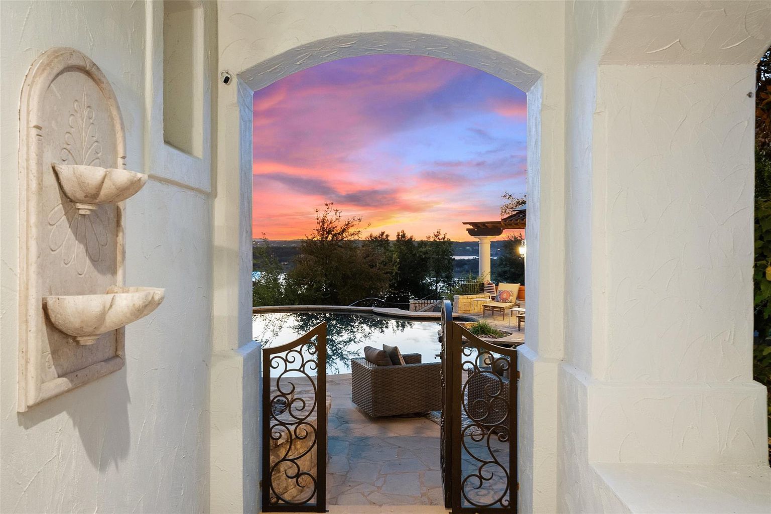 This image showcases a luxurious outdoor living space, viewed through an arched entryway with ornate iron gates. A serene pool reflects the vibrant sunset sky, while comfortable patio furniture invites relaxation. The scene exudes tranquility and upscale living, perfect for entertaining or unwinding.
