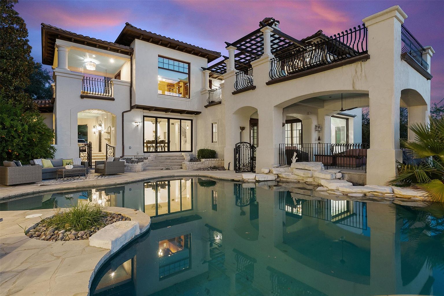 This image showcases the rear exterior of a luxurious home, emphasizing its stunning pool and outdoor living spaces. The architecture features a blend of Mediterranean and modern styles, with white stucco walls, dark-framed windows, and wrought-iron balconies. The pool reflects the home's facade and the colorful sky, creating a serene and inviting atmosphere.