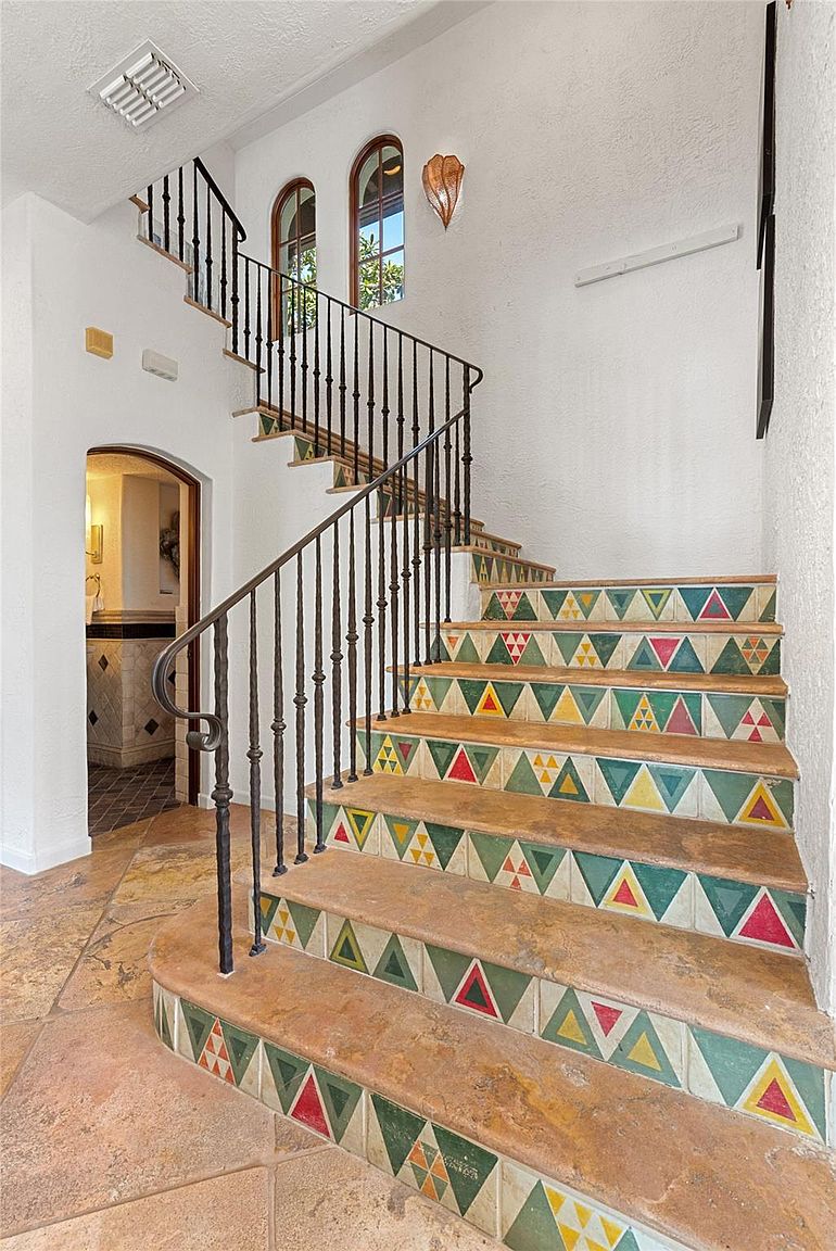 This interior shot showcases a staircase with uniquely patterned tile risers, adding a vibrant and artistic touch to the home. The wrought iron railing complements the warm-toned tile flooring, while arched windows provide natural light. An arched doorway leads to another room, enhancing the home's architectural character.