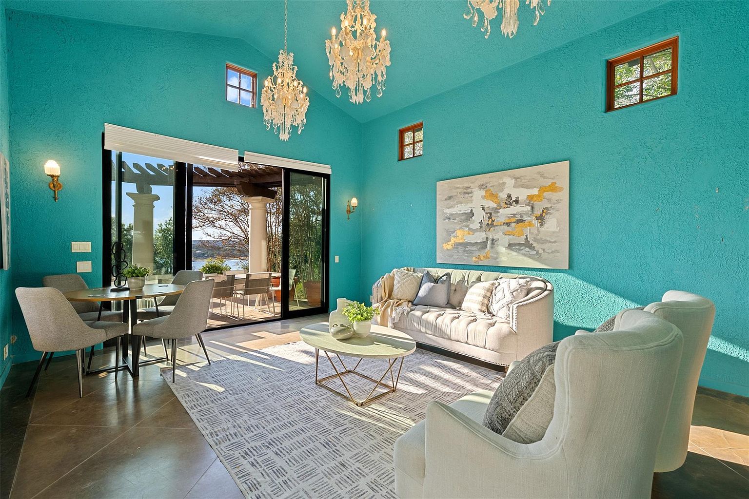 This is an interior shot of a living room featuring a turquoise color scheme. The room is well-lit with natural light coming through large sliding glass doors that lead to an outdoor patio area. The room includes a sofa, two armchairs, a coffee table, a dining table with chairs, and an abstract painting on the wall. Chandeliers hang from the ceiling, adding a touch of elegance.