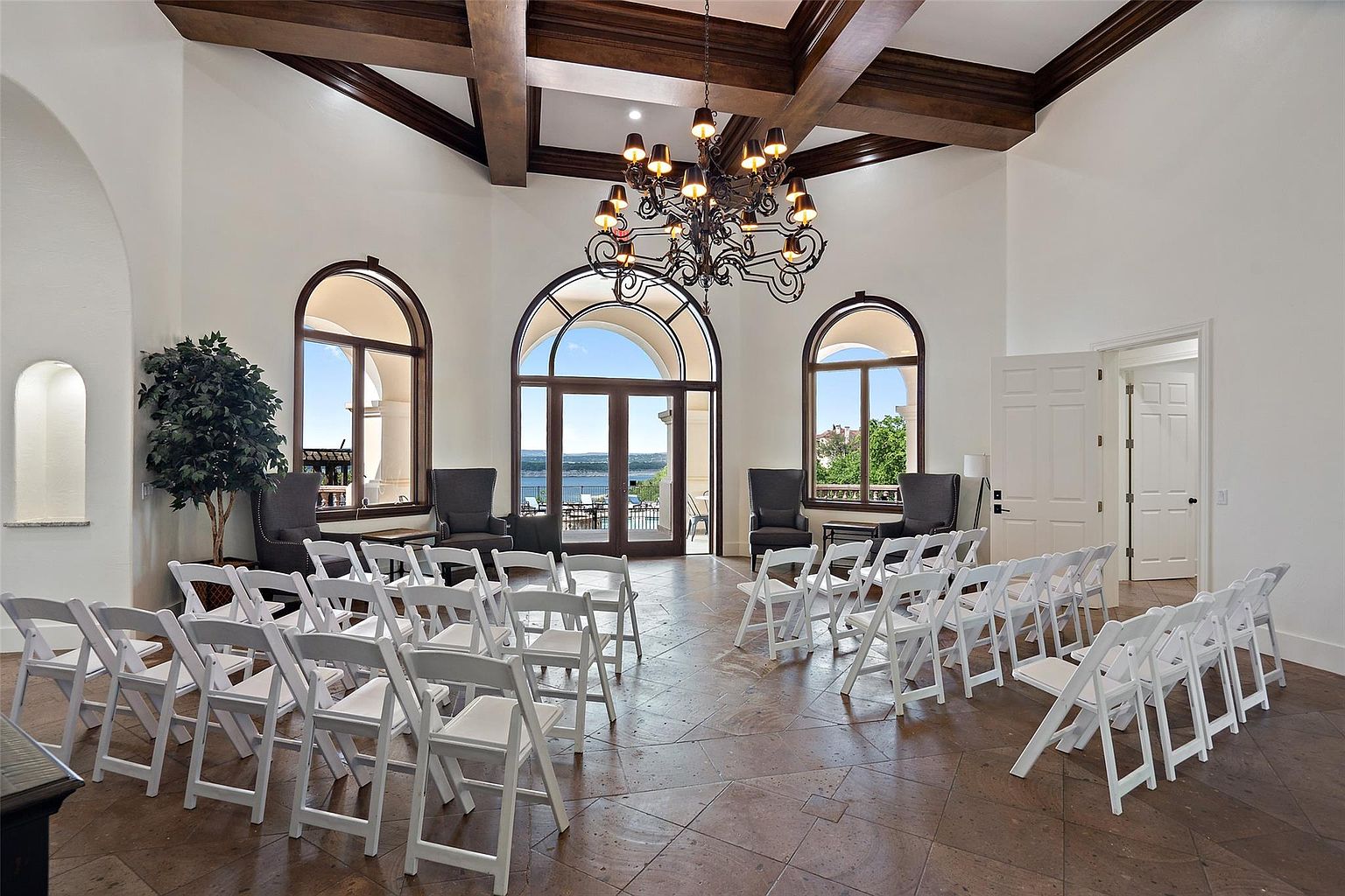 This is an interior shot of a grand living room, likely staged for an event such as a wedding. The room features high ceilings with exposed wooden beams, arched windows offering a view of a lake, and a large ornate chandelier. Rows of white folding chairs are arranged facing the windows, suggesting a ceremony setup.