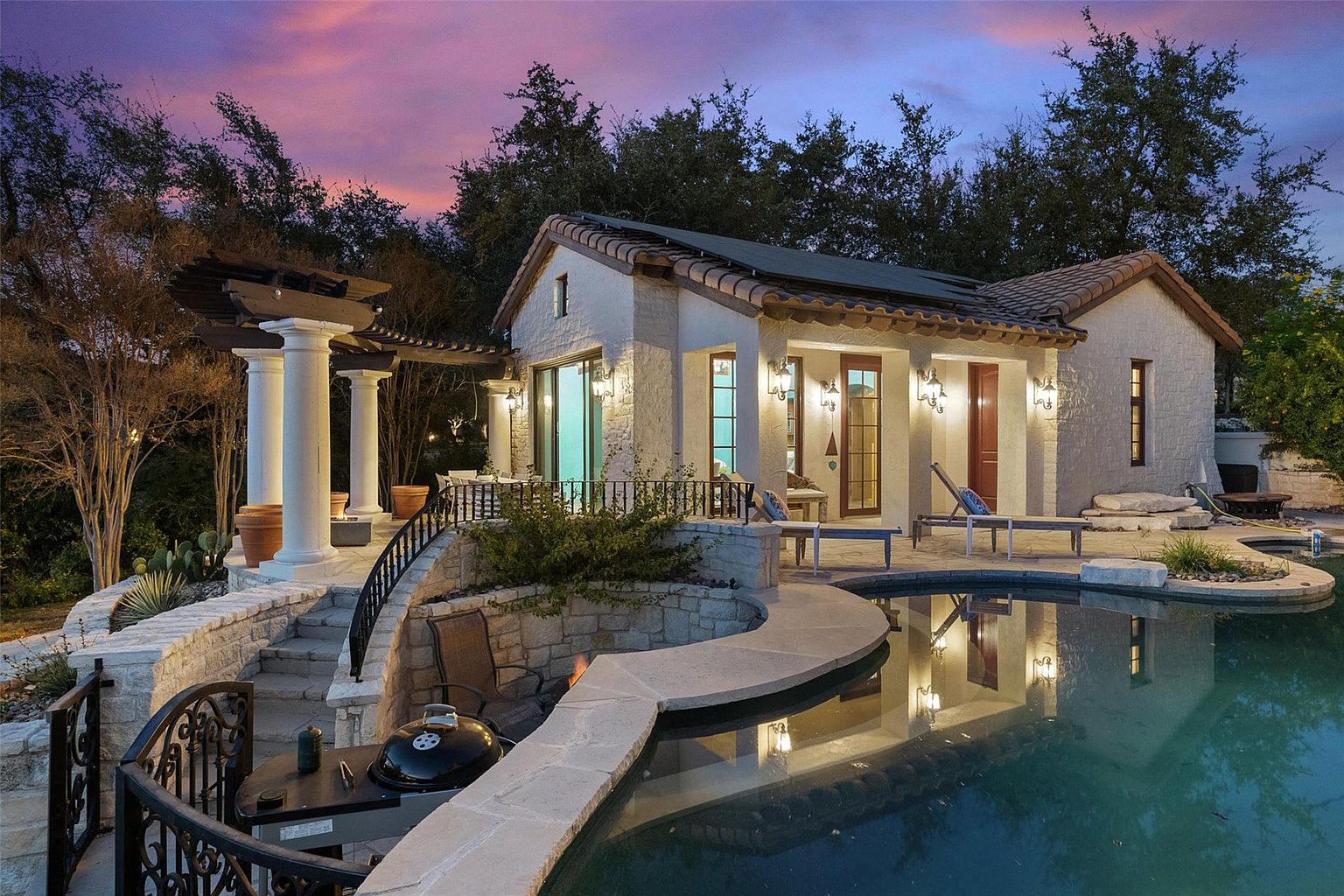 This image showcases a luxurious backyard pool and spa area at dusk. The pool's edge is lined with stone, and the water reflects the warm lights of the adjacent building, which features a tiled roof and stucco walls. A pergola with columns adds an elegant touch, while a built-in grill area suggests outdoor entertaining.