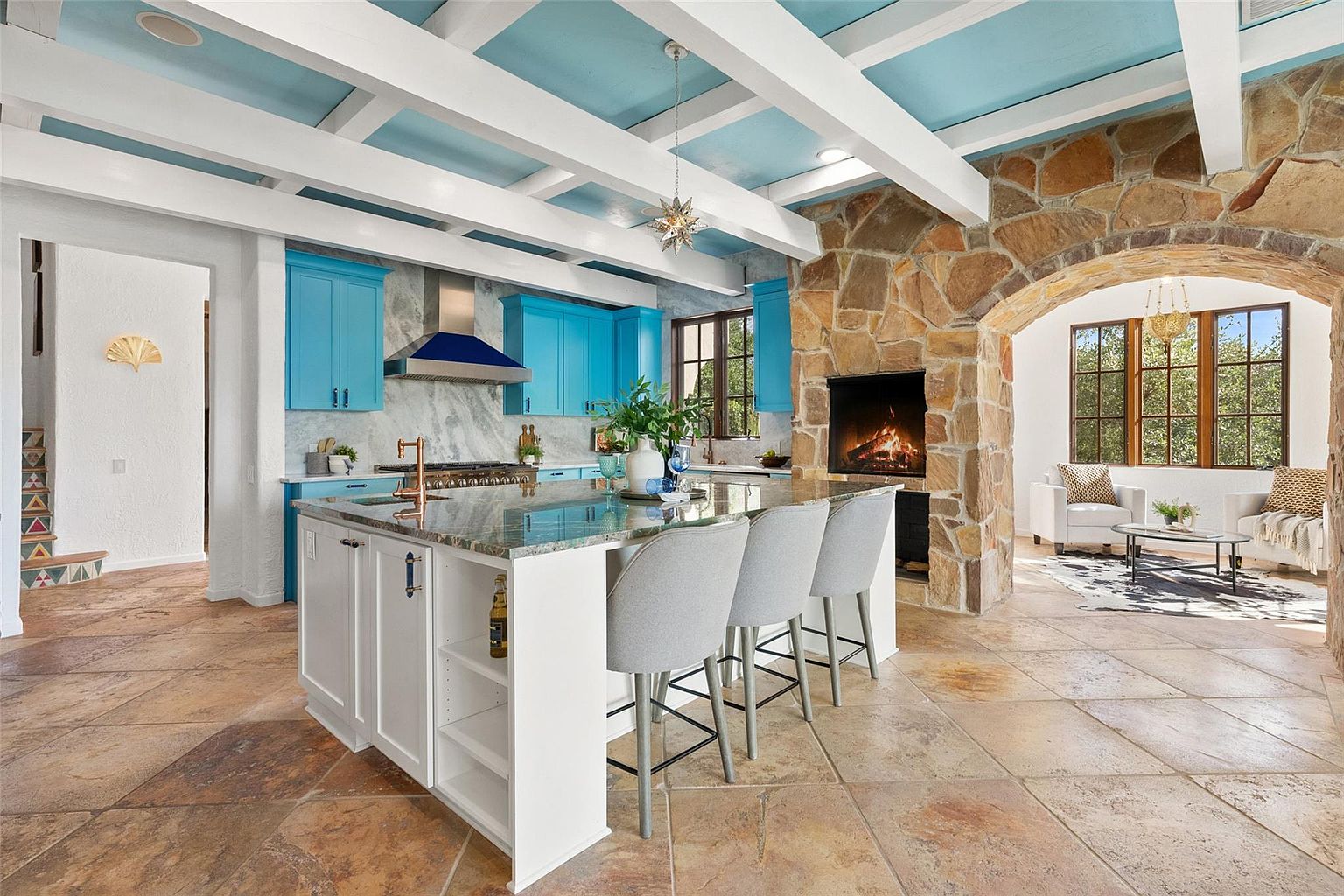 This interior shot showcases a spacious kitchen with a large central island, complete with bar seating and storage. The kitchen features vibrant blue cabinetry, a stone fireplace, and an open layout that flows into a living area. The design blends rustic elements with modern touches, creating a warm and inviting atmosphere.