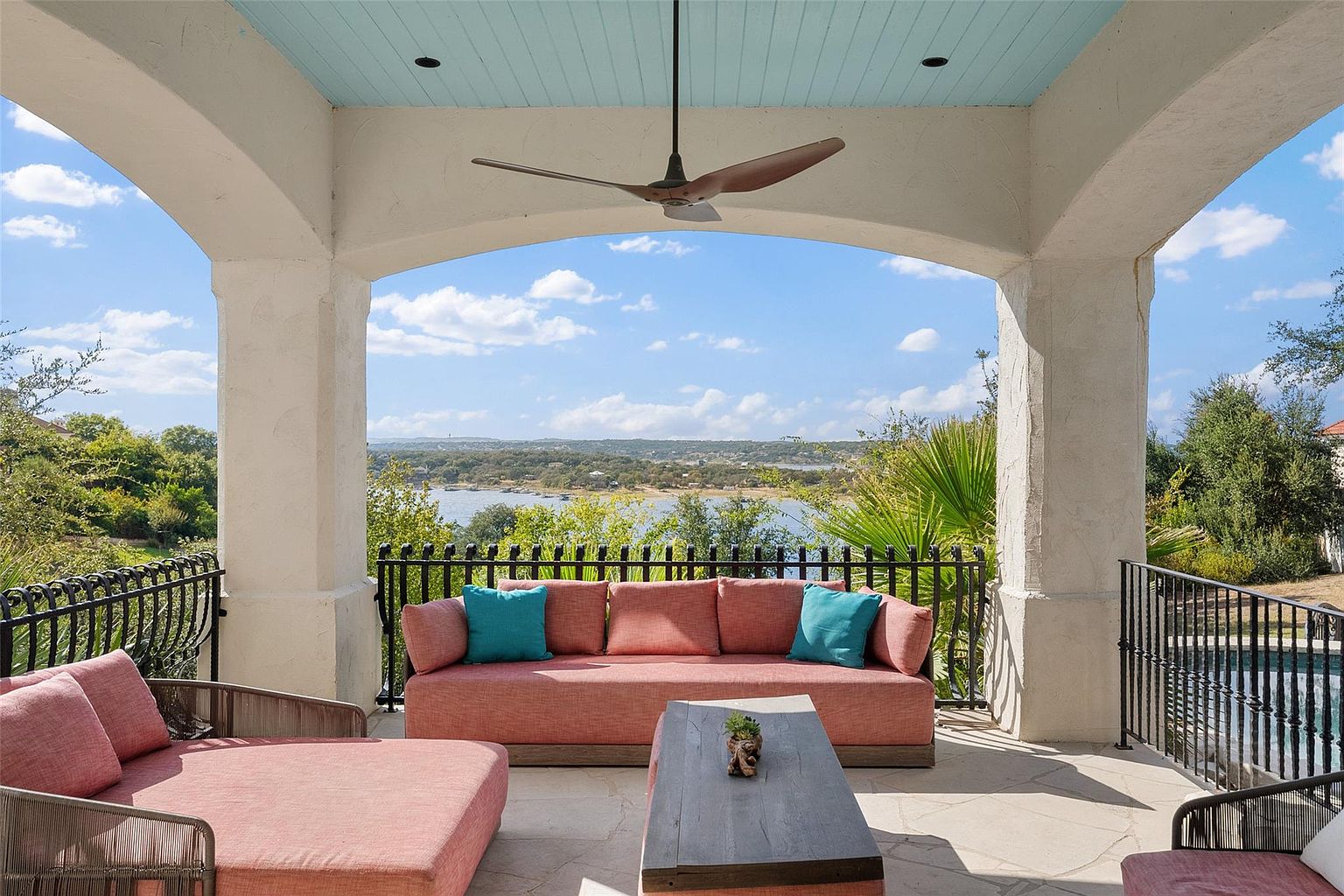 This image showcases a luxurious outdoor living space, specifically a covered patio or balcony. The area is furnished with comfortable seating, including a plush sofa and lounge chairs, all in a coordinating color scheme. A ceiling fan provides air circulation, and the space offers stunning views of a lake or body of water, enhancing the property's appeal for relaxation and entertainment.