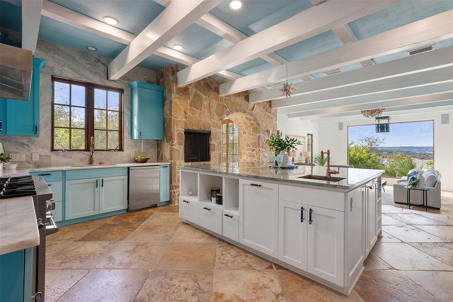 This is a spacious kitchen featuring a large central island with white cabinetry and a contrasting countertop. The kitchen also includes blue cabinets, stainless steel appliances, and a stone accent wall with a fireplace. The room is well-lit with natural light from a large window and recessed lighting, creating a warm and inviting atmosphere.