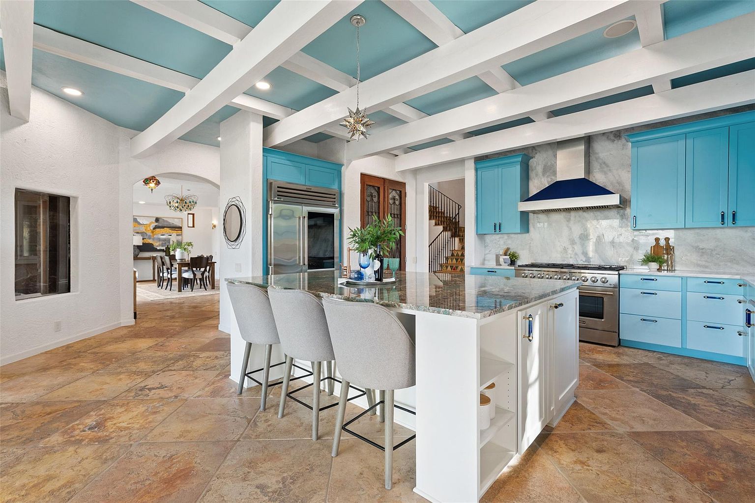 This is a spacious kitchen featuring a large island with seating, blue cabinetry, and stainless steel appliances. The ceiling has exposed white beams and is painted a light blue, adding a unique touch. The flooring is tile, and there is an open view to the dining area and stairs, creating an inviting and connected living space.