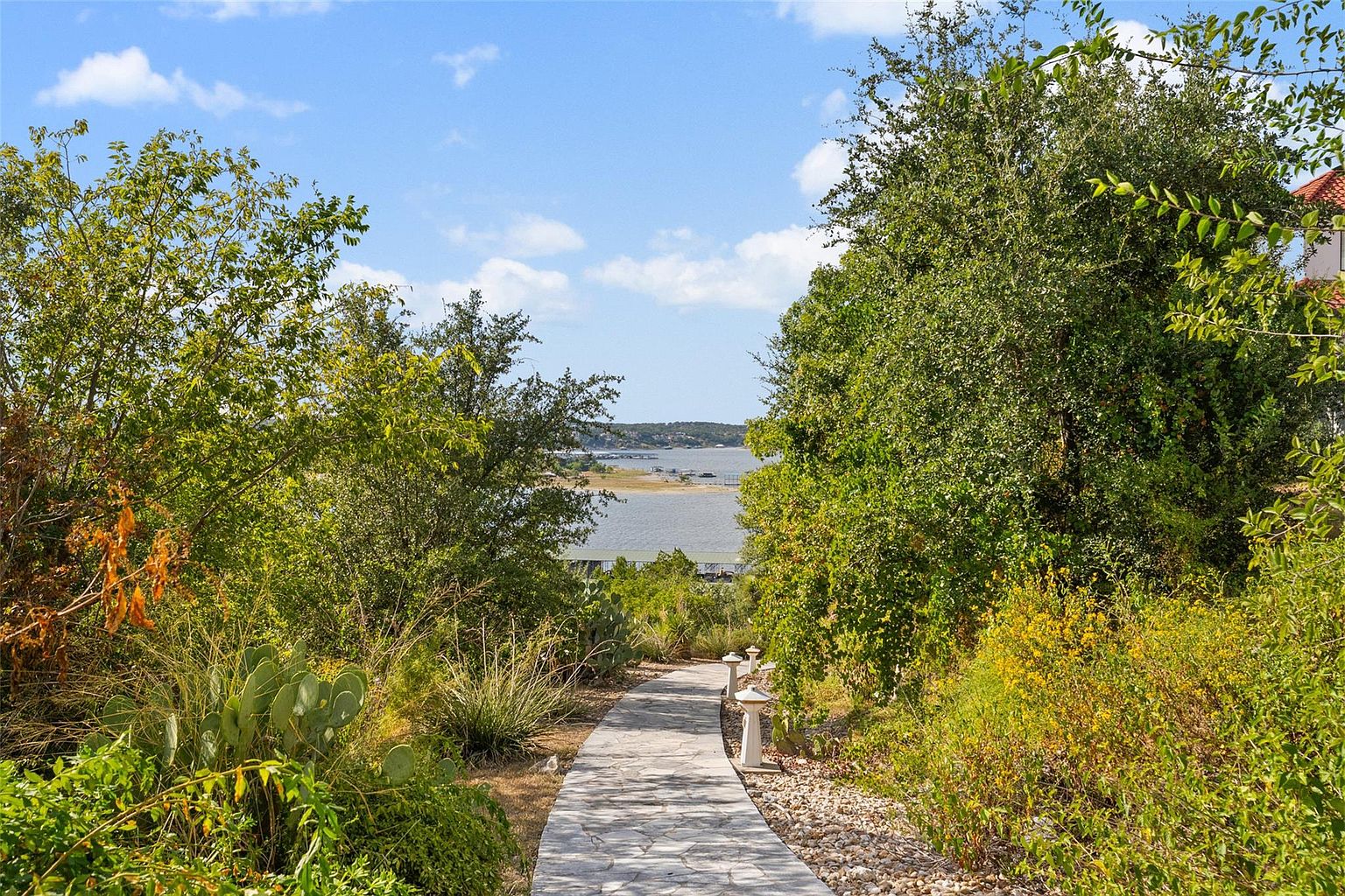 This image showcases a beautifully landscaped pathway leading towards a serene lake view. The stone pathway is flanked by lush greenery, including trees, bushes, and various plants, creating a tranquil and inviting atmosphere. Decorative white lanterns line the path, adding a touch of elegance and guiding the way to the waterfront.