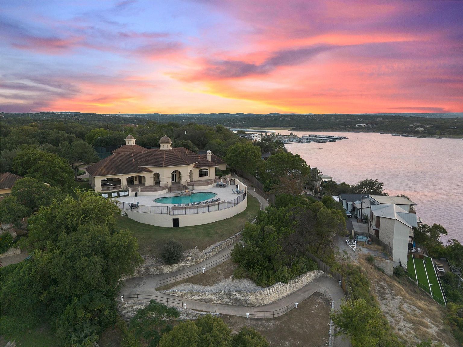 This aerial view showcases a luxurious property with a Mediterranean-style villa featuring a terracotta roof, arched doorways, and a central courtyard with a pool. The villa is situated on a bluff overlooking a lake, with lush landscaping and winding pathways leading to the waterfront. The image captures a serene and upscale living environment at sunset.