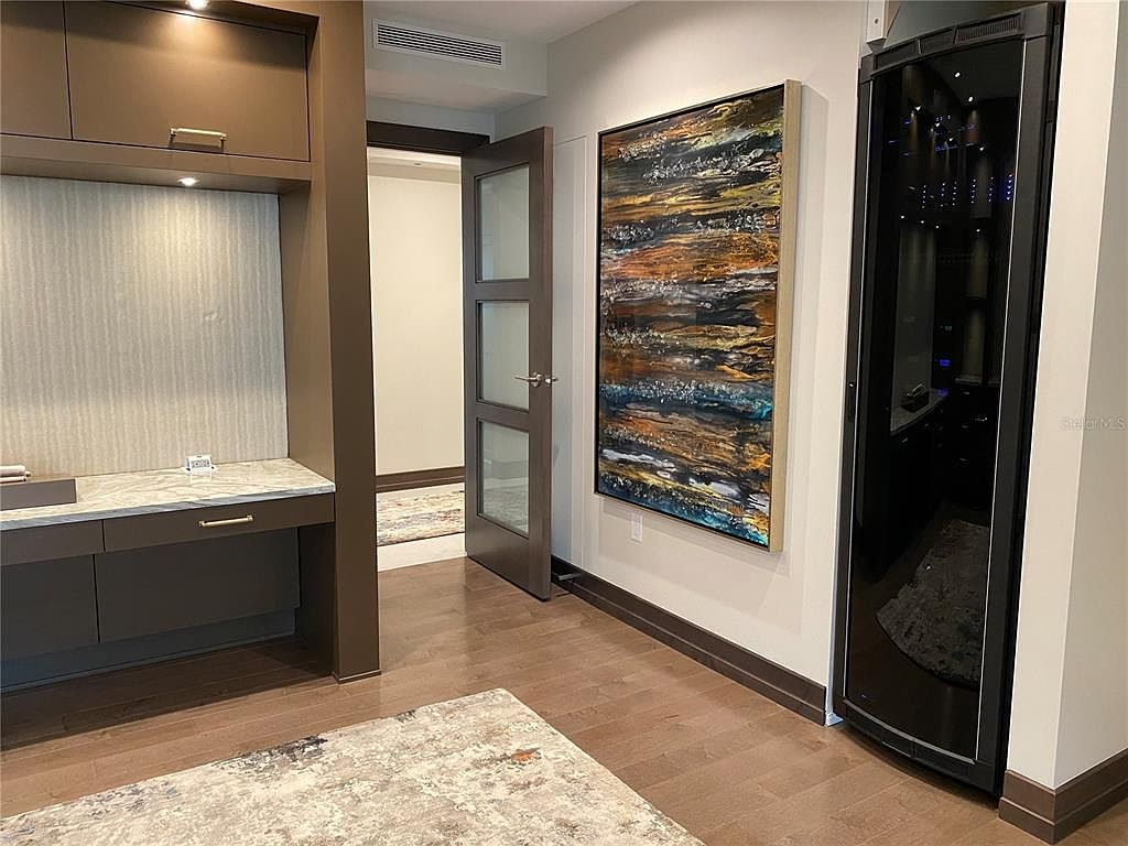 This interior shot showcases a modern office or study area featuring a built-in desk with drawers and upper cabinets in a dark brown finish. An abstract painting hangs on the wall next to a sleek, black wine refrigerator. The room is finished with hardwood flooring and a neutral-toned rug, creating a sophisticated and functional workspace.