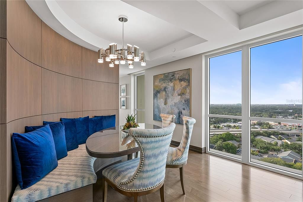 This is an interior shot of a dining room featuring a curved banquette seating area with blue pillows, complemented by stylish chairs around a round table. A modern chandelier hangs above, and large windows offer a panoramic city view, enhancing the room's luxurious and inviting atmosphere. An abstract painting hangs on the wall.