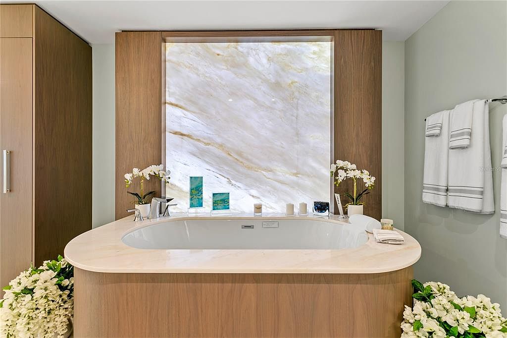 This is a luxurious primary bathroom featuring a large soaking tub with a marble surround and a backlit marble wall panel. The tub is adorned with orchids and decorative items, creating a spa-like atmosphere. The room is finished with light wood cabinetry and soft green walls, complemented by plush white towels.