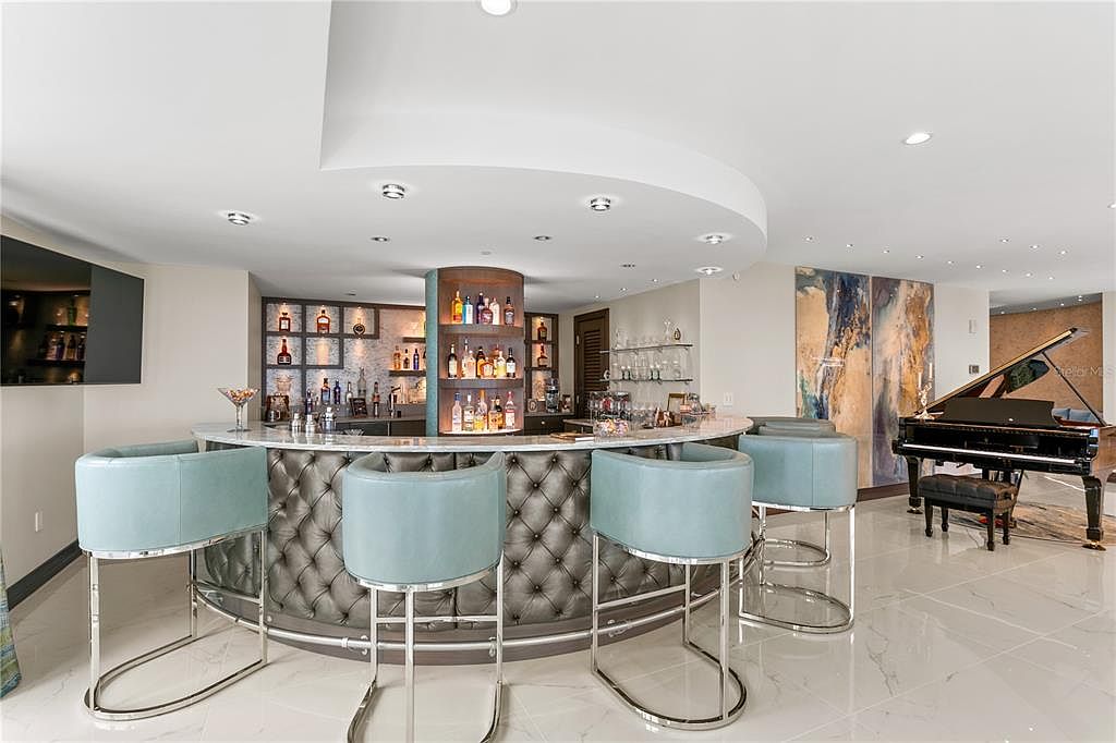 This interior shot showcases a stylish living area featuring a curved bar with tufted detailing and light blue bar stools. A grand piano sits elegantly in the background, complemented by a modern art piece on the wall. The room is well-lit with recessed lighting and features glossy tile flooring, creating a luxurious and inviting atmosphere.