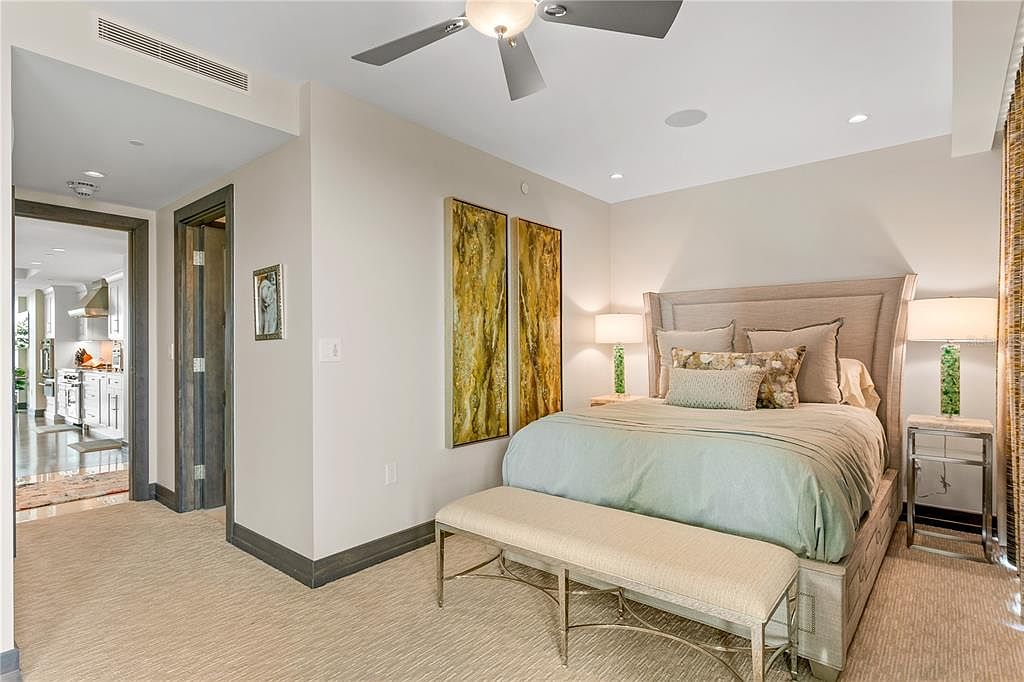 This is a primary bedroom featuring a neutral color palette with a light blue accent on the bedspread. The room includes a large bed with a decorative headboard, two bedside tables with lamps, and a bench at the foot of the bed. Artwork hangs on the wall, and the room is well-lit with recessed lighting and natural light coming through the window.