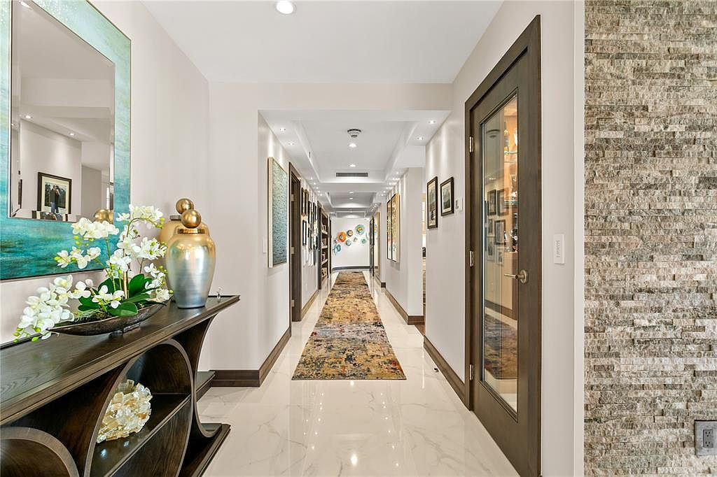 This is an interior shot of a long hallway with marble flooring and recessed lighting. The walls are adorned with artwork, and a decorative console table with floral arrangements sits at the entrance. A patterned runner rug stretches down the center of the hallway, leading the eye towards a distant focal point, and a textured stone wall adds visual interest to the right.