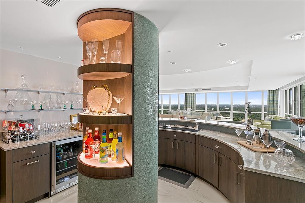 This interior shot showcases a modern kitchen and bar area with custom cabinetry and shelving. The curved bar features a marble countertop and dark wood cabinets, while the adjacent shelving unit displays glassware and liquor bottles. Large windows offer panoramic views, enhancing the luxurious feel of the space.