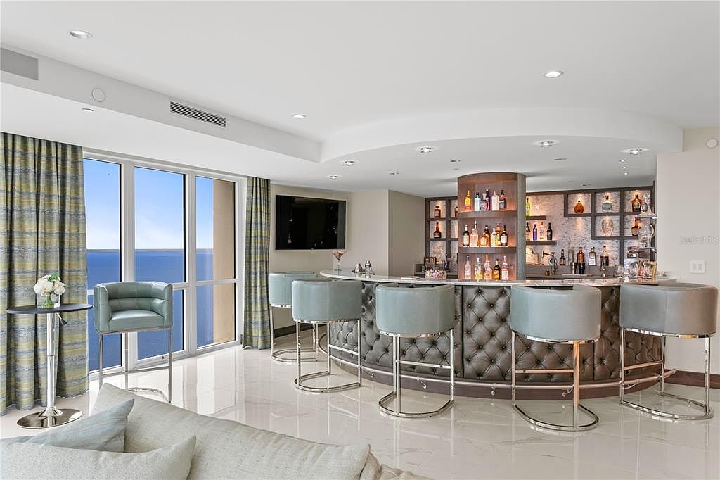 This interior shot showcases a luxurious living room with a custom bar area. The bar features a tufted leather front and a well-stocked display of bottles, complemented by stylish bar stools. Large windows offer a view of the ocean, and the room is furnished with modern seating, creating an elegant and inviting space.