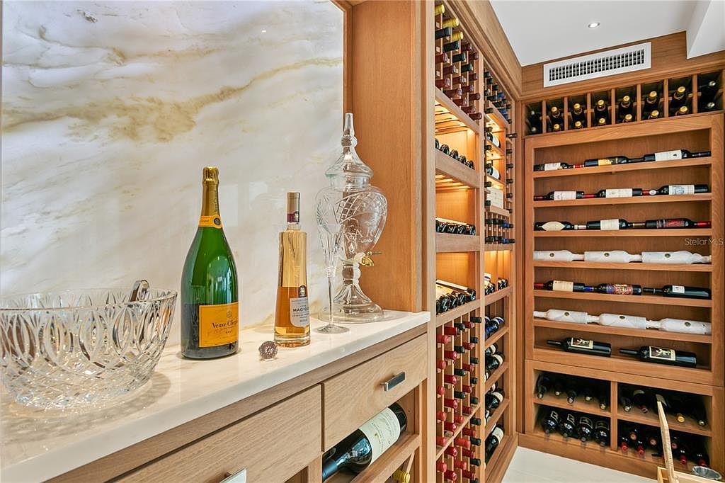 This image showcases a well-organized wine cellar with wooden shelving units filled with numerous wine bottles. A countertop displays decorative items, including a champagne bottle and a crystal ice bucket, adding a touch of elegance. The marble wall and warm lighting create a sophisticated and inviting atmosphere, perfect for wine enthusiasts.