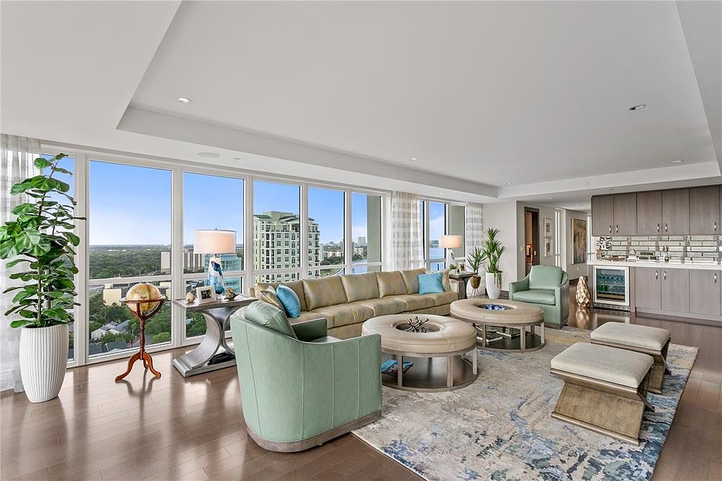 This is a luxurious living room featuring floor-to-ceiling windows with a city view, a large sectional sofa, and two armchairs. The room is decorated with modern furniture, including round coffee tables and stylish ottomans, all set on a patterned area rug. The space is bright and airy, creating an inviting and sophisticated atmosphere.