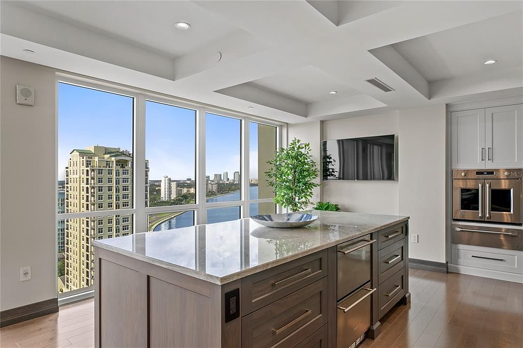 This is a bright and modern kitchen featuring a large island with a marble countertop and dark wood cabinetry. Floor-to-ceiling windows offer a stunning city view, enhancing the luxurious feel of the space. Stainless steel appliances and sleek design elements contribute to the kitchen's contemporary aesthetic.