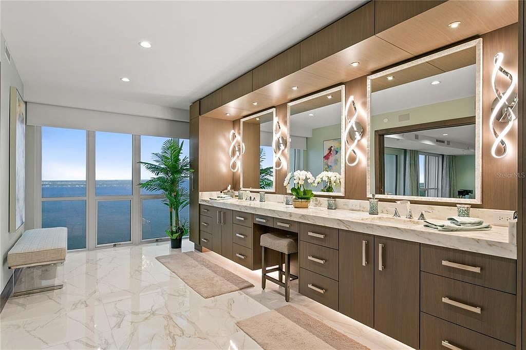 This is a luxurious primary bathroom featuring a long vanity with three mirrors and modern lighting fixtures. The vanity has ample storage with dark wood cabinets and drawers, topped with a light marble countertop. Large windows offer a stunning view, and the marble flooring adds to the elegant and spacious feel of the room.