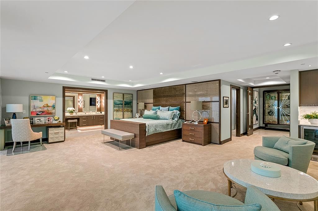 This is a spacious primary bedroom featuring a large bed with a decorative headboard, complemented by matching nightstands. The room includes a sitting area with comfortable armchairs and a coffee table, as well as a desk area. The overall design is luxurious and inviting, with a neutral color palette and ample natural light.