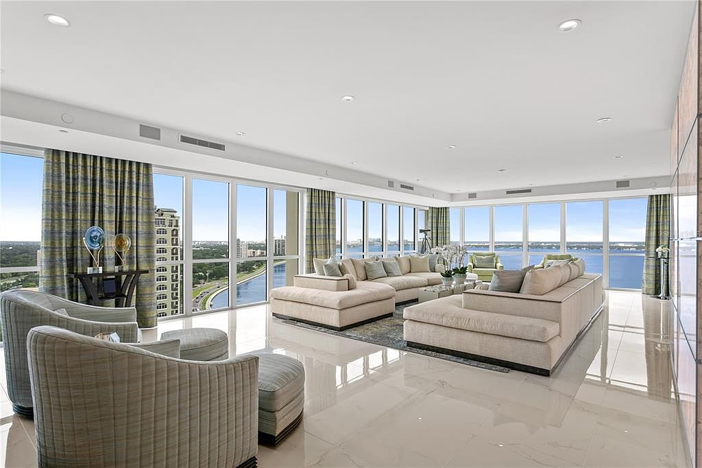 This is a spacious and luxurious living room with floor-to-ceiling windows offering panoramic views of the water and city. The room features a large sectional sofa, two armchairs with ottomans, and a neutral color palette, creating a bright and airy atmosphere. The marble flooring adds to the elegance of the space.