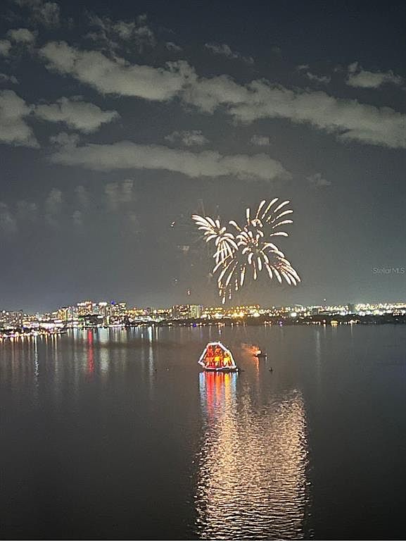 This aerial view captures a stunning fireworks display over a body of water, with a brightly lit structure floating on the water's surface. The city skyline is visible in the background, illuminated with lights, creating a vibrant and festive atmosphere. The scene suggests a waterfront property with access to community events and scenic views.