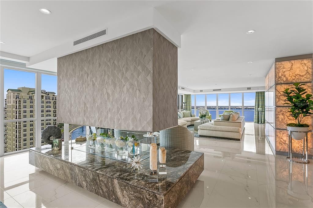This is a luxurious living room featuring a modern fireplace with a marble base and a textured wall. The room has floor-to-ceiling windows offering panoramic views of the water and city. The decor is elegant with neutral tones and plush seating, creating a sophisticated and inviting atmosphere.