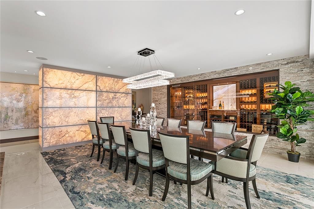 This is an interior shot of a luxurious dining room. The room features a large dining table with seating for ten, a modern chandelier, and a wine cellar built into the wall. The walls are decorated with stone and marble panels, and a large rug covers the floor, creating an elegant and sophisticated atmosphere.