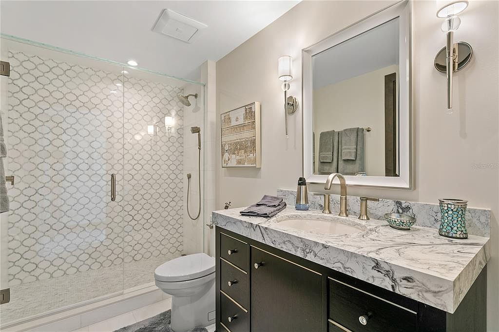 This is a well-lit bathroom featuring a glass-enclosed shower with patterned tile, a toilet, and a vanity with a marble countertop. The vanity has dark wood cabinetry and is complemented by modern lighting fixtures and a decorative mirror. The overall impression is clean, stylish, and functional.