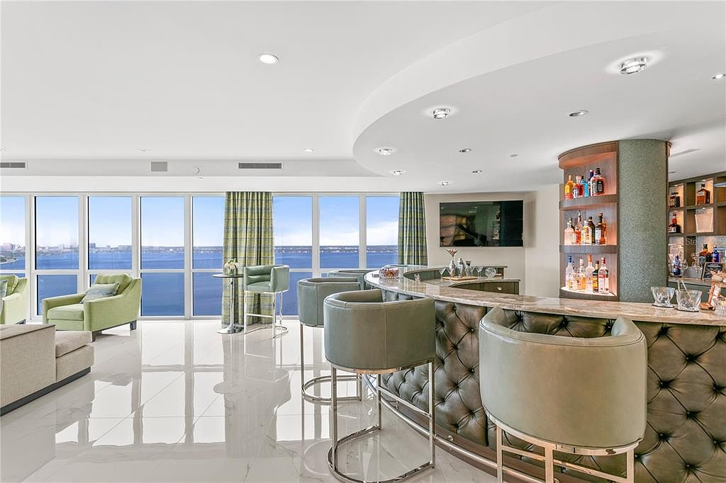 This is an interior shot of a luxurious living room featuring a modern bar area. The room boasts floor-to-ceiling windows offering a stunning water view, complemented by stylish green armchairs and bar stools. The bar area is well-stocked and features a tufted leather front, adding a touch of sophistication to the space.