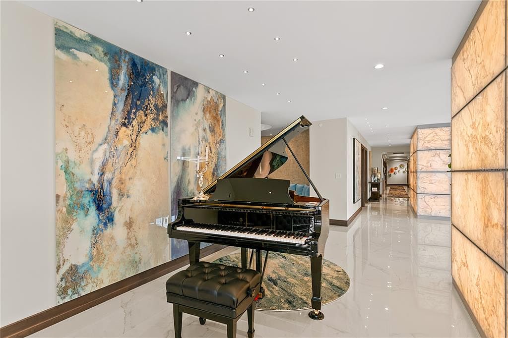 This interior shot showcases a luxurious hallway featuring a grand piano with a black leather bench, complemented by a striking abstract art piece on the wall. The hallway is illuminated by recessed lighting and features polished marble flooring, leading to a corridor with decorative wall panels. The overall impression is one of elegance and sophistication.