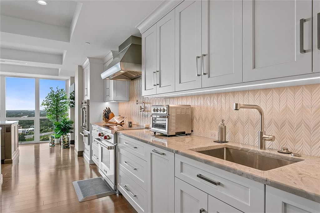 This is a bright and modern kitchen featuring white cabinetry with sleek hardware, stainless steel appliances, and light-colored countertops. The backsplash has a unique leaf-like pattern, and a large window provides natural light and a view of the outdoors. The perspective is from a medium distance, showcasing the kitchen's layout and design.