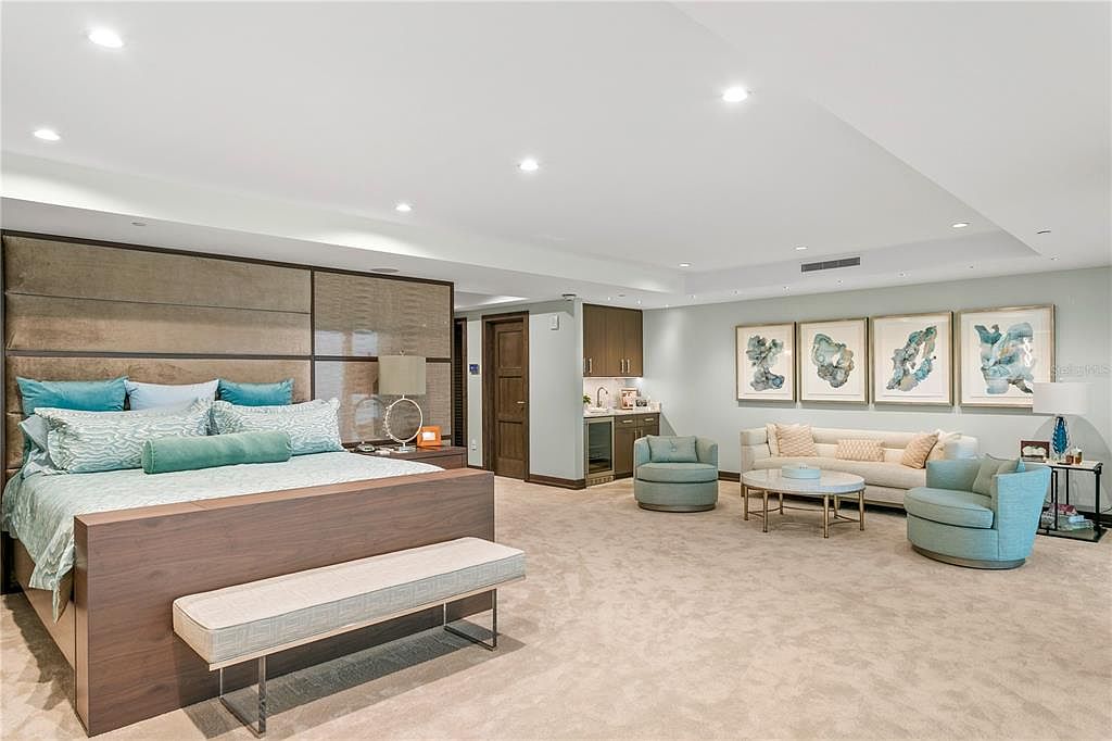 This is a spacious primary bedroom featuring a large bed with a padded headboard and a seating area. The room is carpeted and has recessed lighting. The color palette is soft and calming, creating a luxurious and relaxing atmosphere.