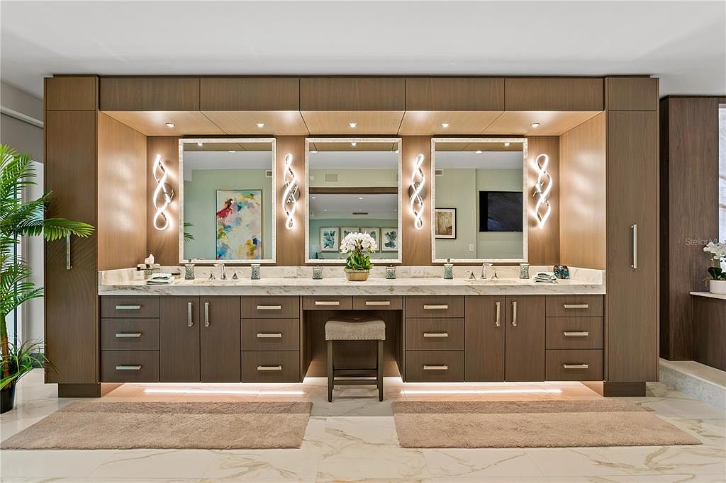 This is a well-lit primary bathroom featuring a double vanity with marble countertops and dark wood cabinetry. Three mirrors with decorative lighting fixtures hang above the sinks, and a makeup vanity with a stool is centered between the two sink areas. The flooring is marble, and there are matching rugs in front of each sink area, creating a luxurious and functional space.