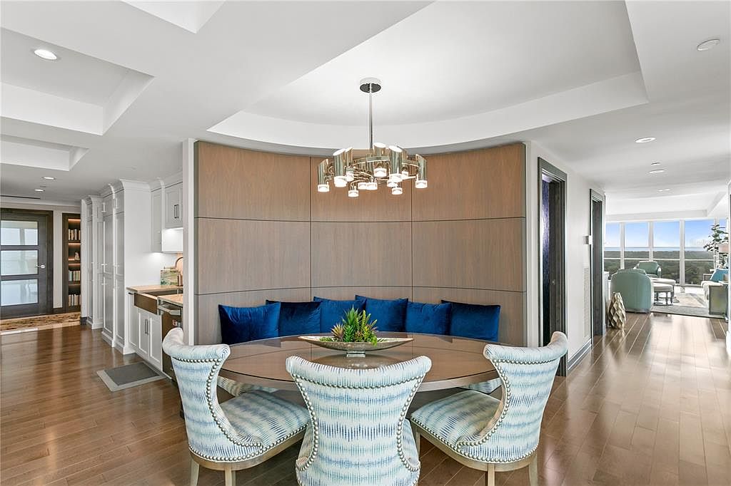 This is an interior shot of a dining room featuring a round table with upholstered chairs and a built-in banquette with blue cushions. A modern chandelier hangs above the table, and the room has hardwood floors. The space is well-lit and appears to be part of a luxurious home.