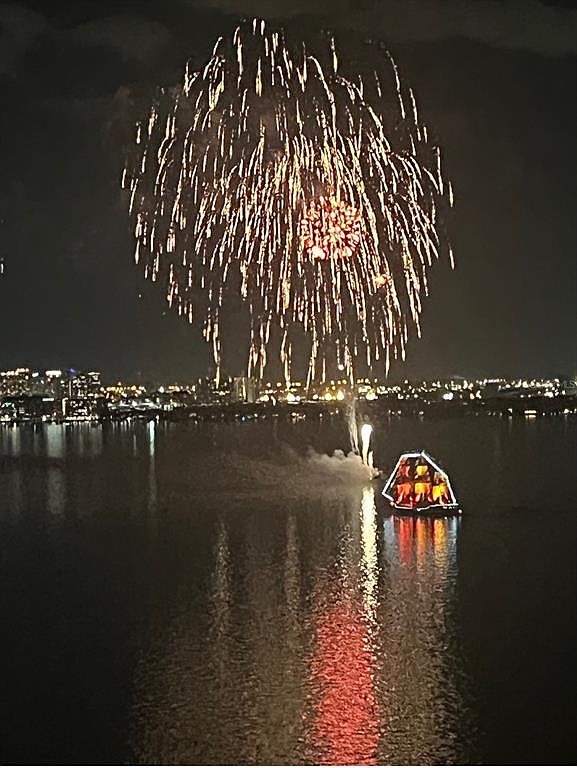 This image showcases a vibrant fireworks display over a body of water, with city lights visible in the background. A structure illuminated with warm light sits on the water, reflecting in the dark surface. The scene creates a festive and visually striking atmosphere, potentially highlighting the location's appeal for events and entertainment.