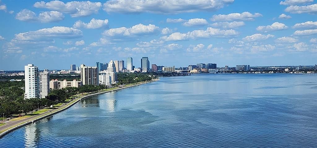 This aerial view showcases a stunning cityscape along a waterfront. The image features a mix of high-rise buildings and lush greenery, with a clear blue sky dotted with fluffy clouds above. The calm water reflects the sky and buildings, creating a serene and picturesque scene, ideal for highlighting the location's beauty and urban appeal.