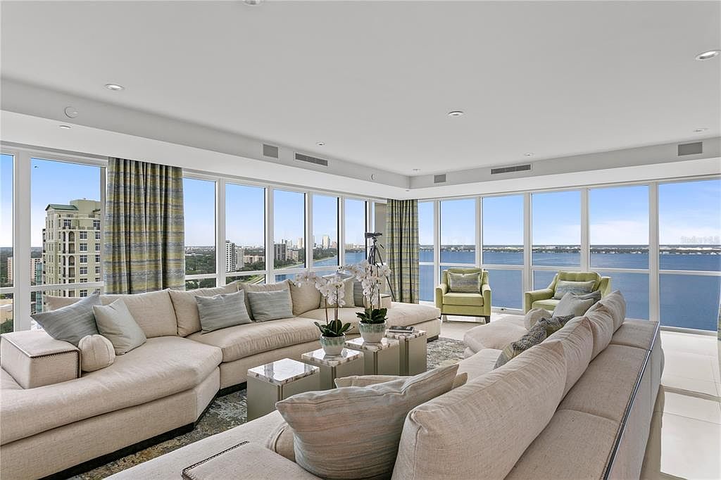 This is a luxurious living room with floor-to-ceiling windows offering panoramic water views. The room features two large, comfortable sectional sofas with neutral tones and decorative pillows. Two light green armchairs and two marble-topped side tables with orchids add to the elegant ambiance, creating a sophisticated and inviting space.
