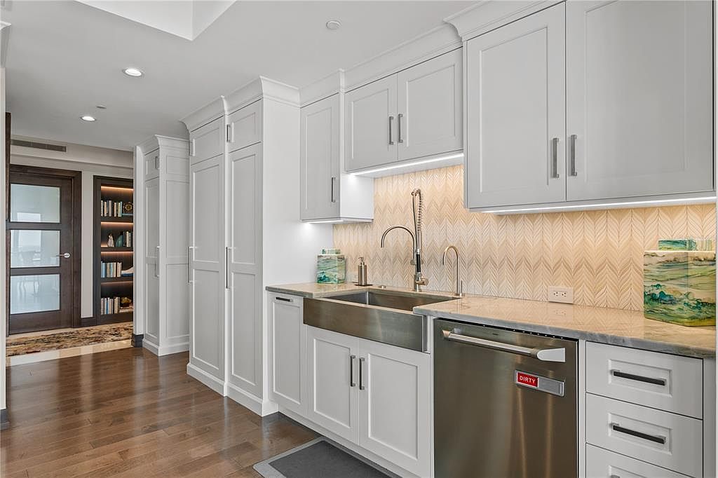 This is a well-lit kitchen featuring white cabinetry with sleek, modern hardware. A stainless steel farmhouse sink and appliances complement the light countertops and patterned backsplash. The hardwood flooring adds warmth to the space, and the overall design is clean and sophisticated.
