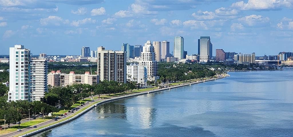 This aerial view showcases a stunning waterfront cityscape with a mix of high-rise residential buildings and commercial skyscrapers. The coastline is lined with lush greenery and a scenic road, creating a harmonious blend of urban and natural elements. The calm, reflective water adds to the overall sense of tranquility and luxury.