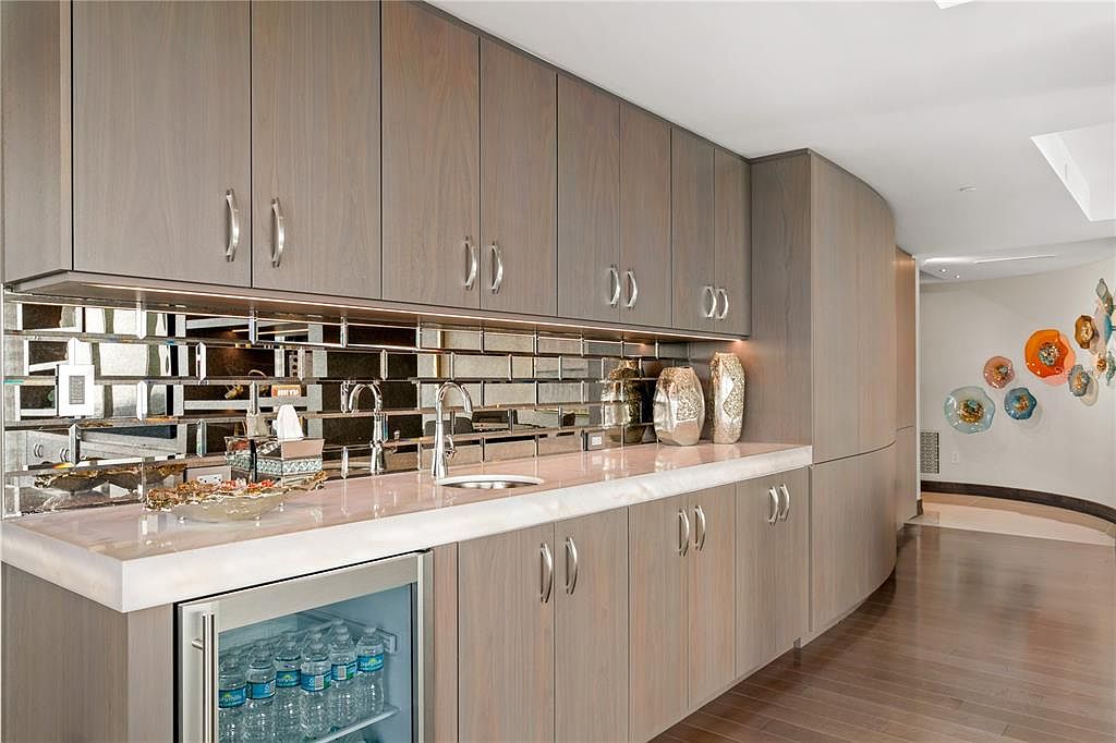 This image showcases a modern kitchen area with sleek, light wood cabinetry and silver hardware. A mirrored backsplash adds depth and reflects light, complementing the light-colored countertop. A built-in beverage refrigerator is visible, and decorative vases add a touch of elegance.