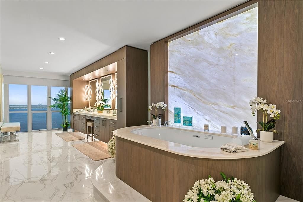 This is a luxurious primary bathroom featuring a large soaking tub with a marble backdrop and wood paneling. The room includes a vanity area with dual sinks, illuminated mirrors, and ample storage. Large windows offer a view of the water, creating a spa-like atmosphere with a clean and modern design.