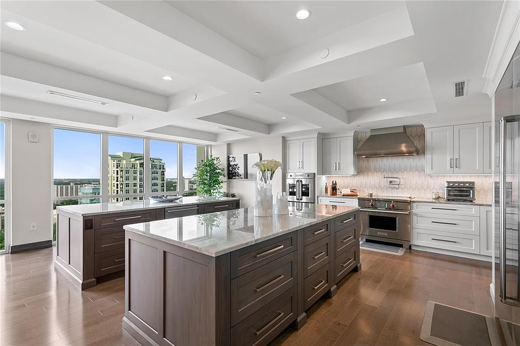 This is a bright and spacious kitchen featuring two large islands with marble countertops and dark wood cabinetry. The kitchen also includes white cabinets, stainless steel appliances, and large windows offering a city view. The overall design is modern and luxurious, creating an inviting space for cooking and entertaining.