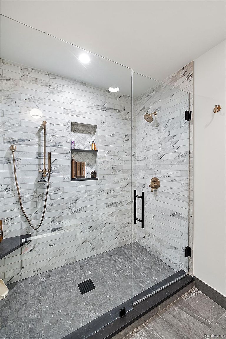 This is a modern primary bathroom featuring a glass-enclosed shower with marble-like tile walls and a gray tiled floor. The shower includes a built-in niche for toiletries and bronze fixtures, adding a touch of elegance. The design is clean and contemporary, creating a spa-like atmosphere.